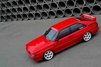 Punainen Audi Ur-Quattro seisoo harmaalla tiellä, takaspoileri ja valkoiset vanteet erottuvat taustalla olevien betoniputkien edessä.